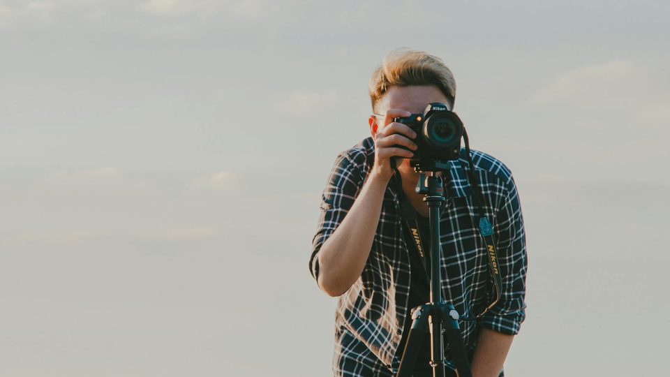 A person holding a camera beside a tripod outdoors.